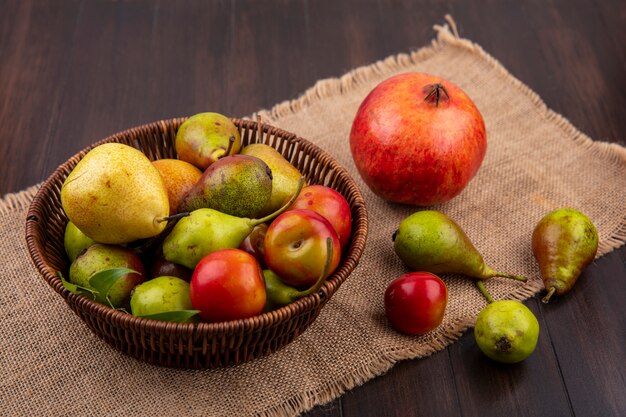 Vista frontal de frutas como durazno manzana ciruela en canasta con granada sobre tela de saco sobre superficie de madera