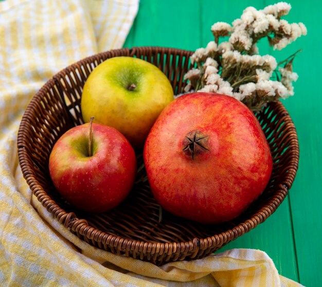 Vista frontal de frutas como manzana y granada con flores en canasta sobre tela escocesa y superficie verde