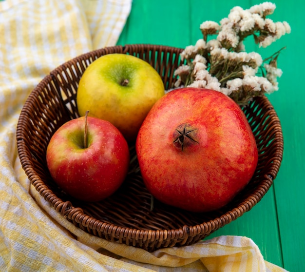 Vista frontal de frutas como manzana y granada con flores en canasta sobre tela escocesa y superficie verde