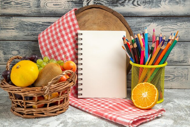 Vista frontal de frutas frescas con bloc de notas y lápices sobre fondo gris color de cuaderno de frutas cítricas