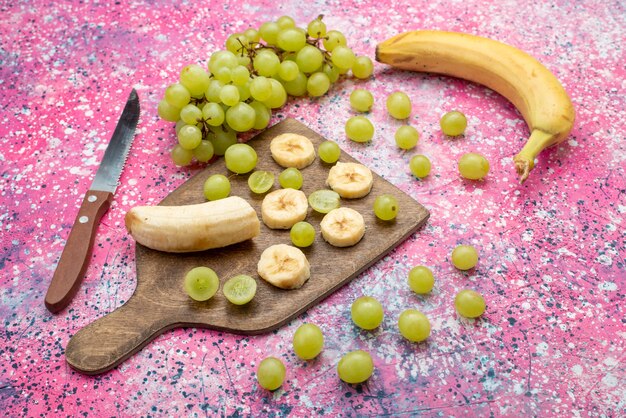 Vista frontal de frutas frescas en rodajas, uvas y plátanos en el escritorio púrpura color de jugo suave de frutas