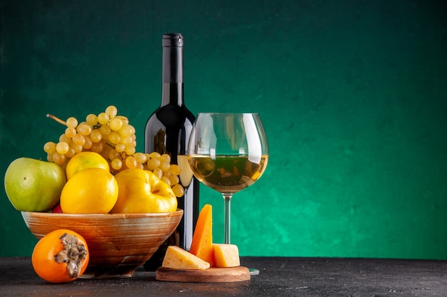 Vista frontal de frutas frescas en un tazón de madera, manzanas, membrillo, limón, uvas, caqui, botella de vino y queso de vidrio sobre tablero de madera en la mesa verde con lugar libre