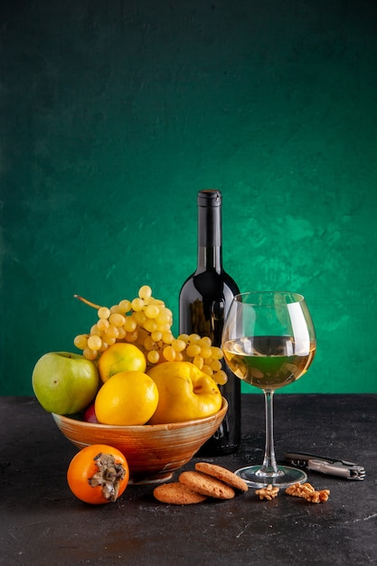 Vista frontal de frutas frescas en un tazón de manzana, membrillo, limón, uvas, caqui, botella de vino y galletas de vidrio, abridor de vino en la mesa verde