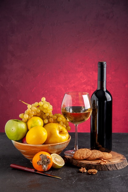 Vista frontal de frutas frescas en un tazón de manzana, membrillo, uvas, caqui, botella de vino, copa de vino, galletas en tablero de madera en la mesa roja