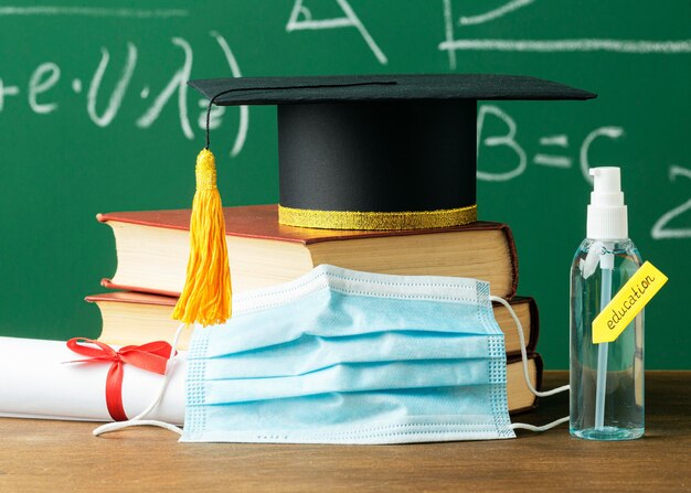 Vista frontal de la gorra académica en libros con máscara médica y desinfectante de manos