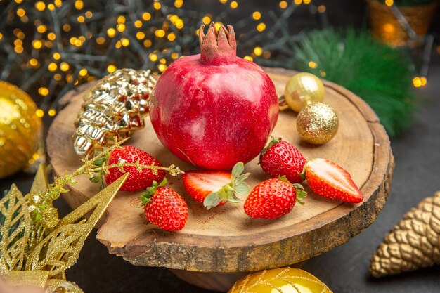 Vista frontal de granada fresca con fresas alrededor de juguetes navideños sobre fondo oscuro, fotografía en color, frutas navideñas navideñas