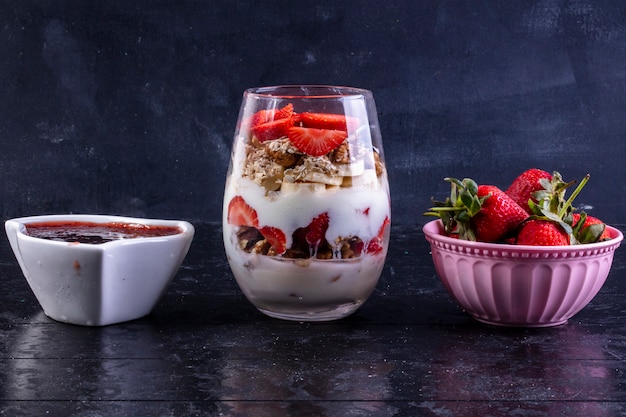 Vista frontal de granola con frutas y nueces en un vaso con fresas en un tazón rosa y mermelada