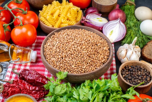 Vista frontal de granos de trigo sarraceno en pastas de toalla despojada roja verduras frescas rebanadas de pan botella de aceite caído un paquete verde sobre mesa negra