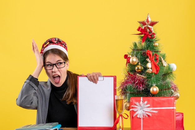 Vista frontal gritando niña con sombrero de Navidad sentado en la mesa árbol de Navidad y cóctel de regalos