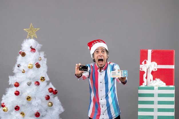 Vista frontal gritó hombre con tarjeta y boleto de viaje mirando alto alrededor del árbol de Navidad y regalos