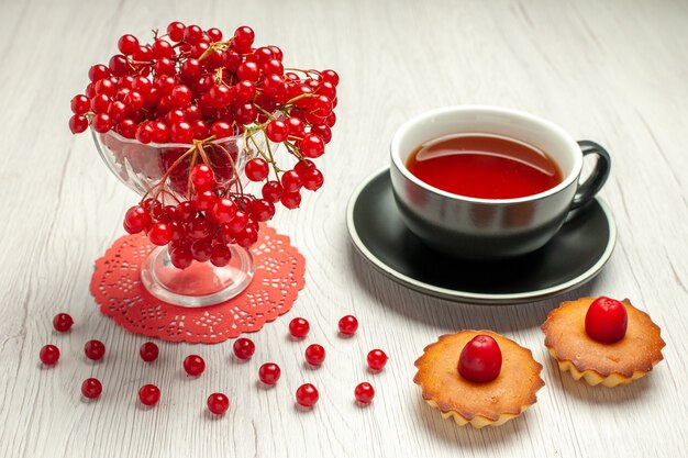 Vista frontal de grosella roja en un vaso de cristal sobre el tapete de encaje ovalado rojo una taza de té y tartas en la mesa de madera blanca