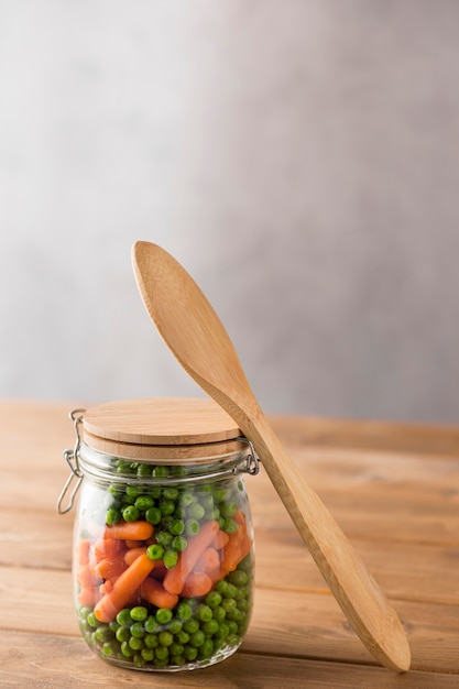 Vista frontal de guisantes y zanahorias pequeñas en frasco de vidrio con cuchara y espacio de copia
