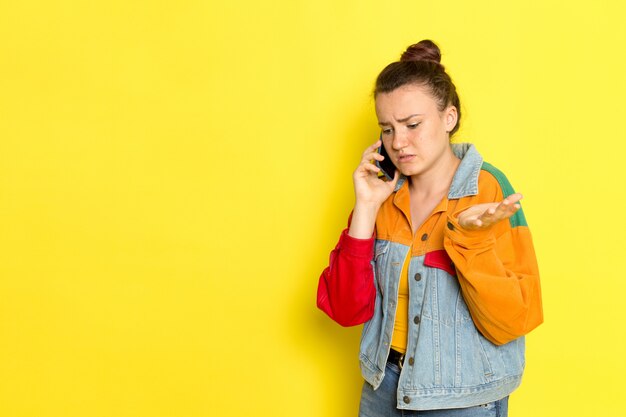 Una vista frontal hembra joven en camisa amarilla chaqueta colorida y jeans azul hablando por teléfono