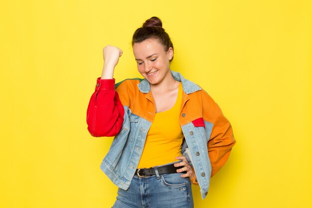 Una vista frontal hembra joven en camisa amarilla chaqueta colorida y jeans azul posando y mostrando su puño con sonrisa