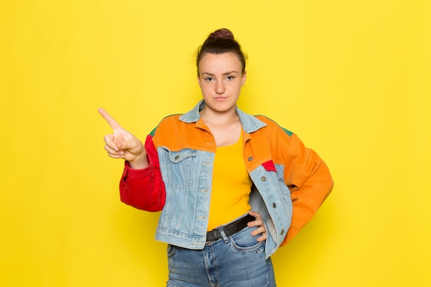 Una vista frontal hembra joven en camisa amarilla chaqueta colorida y jeans azul posando