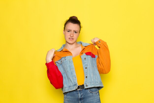 Una vista frontal hembra joven en camisa amarilla chaqueta colorida y jeans azul posando