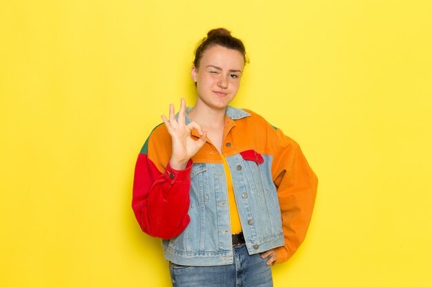 Una vista frontal hembra joven en camisa amarilla chaqueta colorida y jeans azul posando