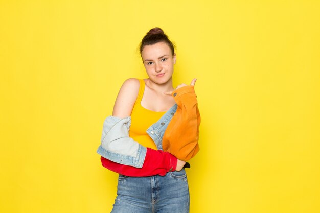 Una vista frontal hembra joven en camisa amarilla chaqueta colorida y jeans azul simplemente posando