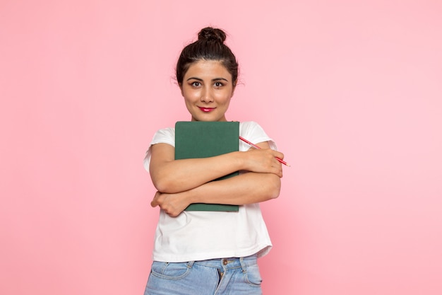 Una vista frontal hembra joven en camiseta blanca y jeans azul sosteniendo un cuaderno con sonrisa