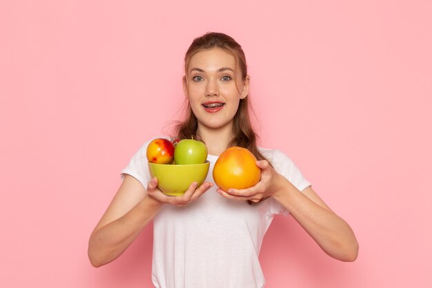 Vista frontal de la hembra joven en camiseta blanca con placa con frutas frescas y pomelo en la pared de luz