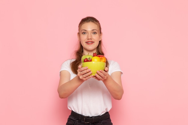 Vista frontal de la hembra joven en camiseta blanca con plato con frutas frescas en la pared rosa claro