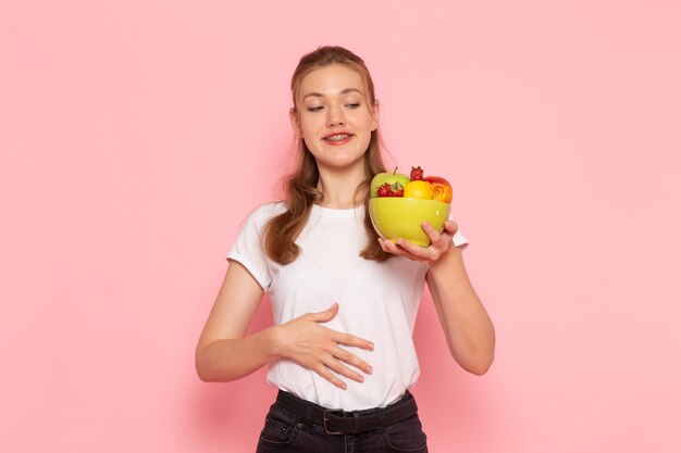 Vista frontal de la hembra joven en camiseta blanca con plato con frutas frescas en la pared rosa claro