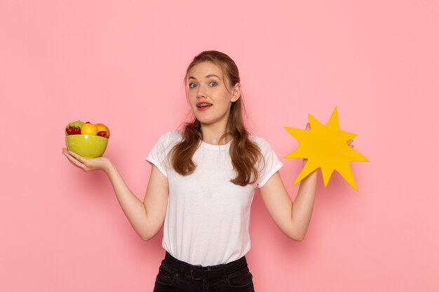 Vista frontal de la hembra joven en camiseta blanca con plato con frutas y gran cartel amarillo en la pared rosa claro