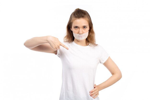 Una vista frontal hembra joven en camiseta blanca con vendaje blanco alrededor de su boca, señalando con el dedo en el blanco