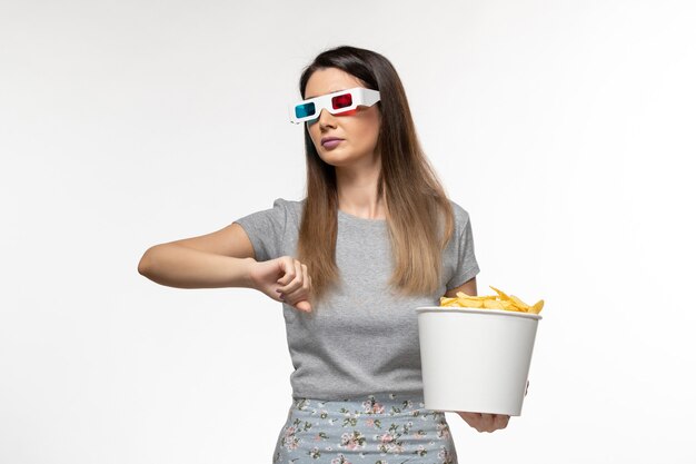 Vista frontal hembra joven comiendo cips mientras ve la película en gafas de sol d mirando su muñeca sobre una superficie blanca