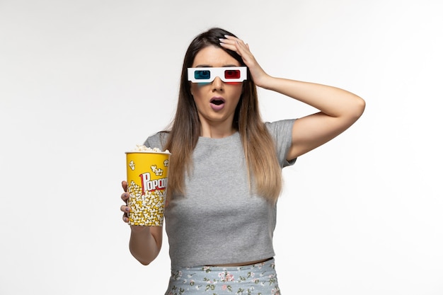 Vista frontal hembra joven comiendo paquete de palomitas de maíz y viendo películas en gafas de sol d sobre superficie blanca