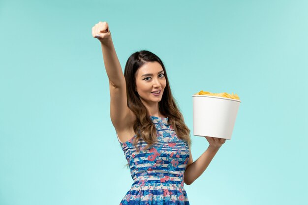 Vista frontal hembra joven sosteniendo la cesta con papas fritas en la superficie azul