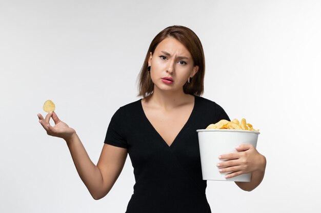 Vista frontal hembra joven sosteniendo la cesta con patatas fritas y comiendo en la superficie blanca