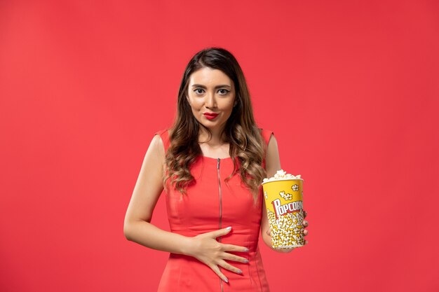 Vista frontal hembra joven sosteniendo el paquete de palomitas de maíz en la superficie de color rojo claro