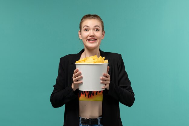 Vista frontal hembra joven sosteniendo el paquete con papas fritas y viendo una película en la superficie azul