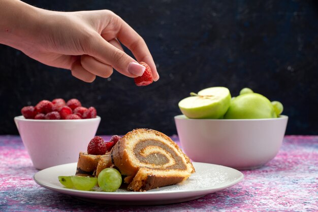 Vista frontal hembra tomando frambuesa de plat ewith roll cake junto con manzanas y frambuesas en la oscuridad