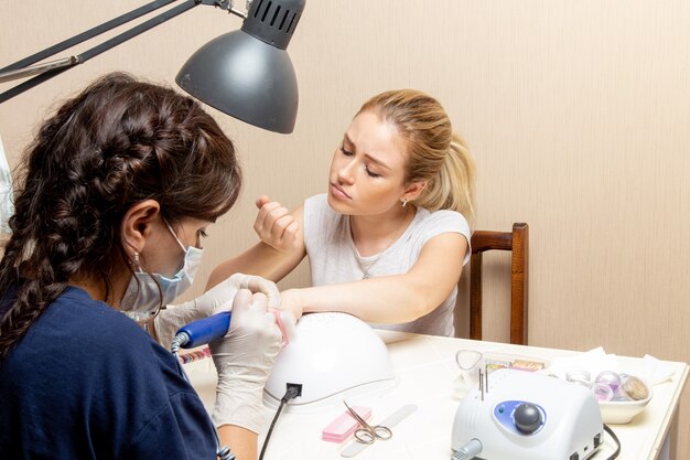 Vista frontal de las hembras jóvenes arreglando sus uñas por manicurista dentro de la habitación belleza mujer manicura uñas autocuidado dama