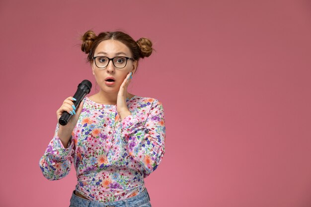 Vista frontal de las hembras jóvenes en flor diseñada camisa y jeans sosteniendo el micrófono tratando de cantar sobre el fondo rosa