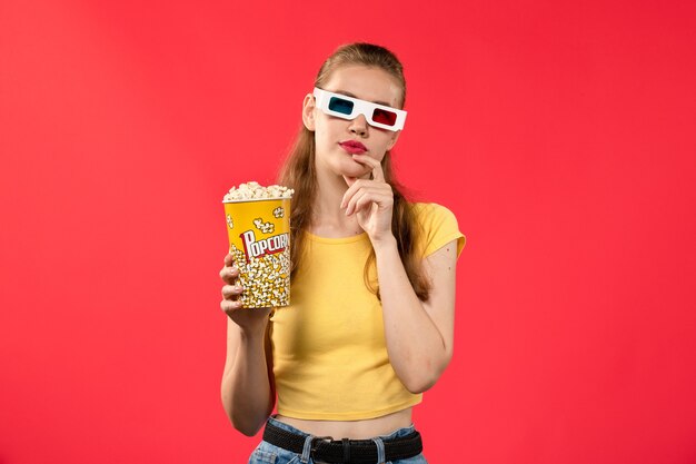 Vista frontal de las hembras jóvenes que sostienen el paquete de palomitas de maíz en gafas de sol d en la pared de color rojo claro películas de cine