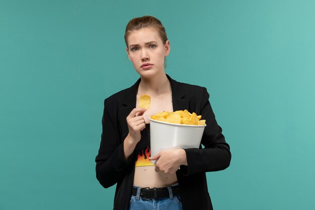 Vista frontal de las hembras jóvenes sosteniendo y comiendo patatas fritas viendo la película en la superficie azul claro