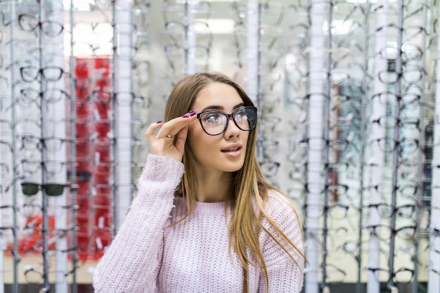 Vista frontal de la hermosa chica en suéter blanco pruebe gafas en tienda profesional en