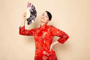Foto gratuita una vista frontal hermosa geisha japonesa en tradicional vestido japonés rojo con palos de pelo posando sosteniendo abanico plegable sonriendo sobre la ceremonia de fondo crema japón