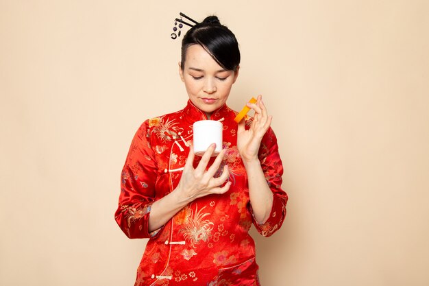 Una vista frontal hermosa geisha japonesa en el tradicional vestido rojo japonés con bastones para el cabello posando lata de crema de apertura en la ceremonia de fondo crema Japón