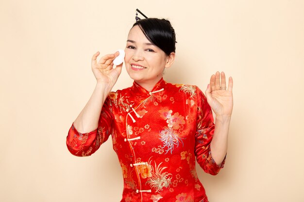 Una vista frontal hermosa geisha japonesa en el tradicional vestido rojo japonés con bastones para el cabello posando sosteniendo un poco de algodón blanco limpiando su rostro en la ceremonia de fondo crema Japón