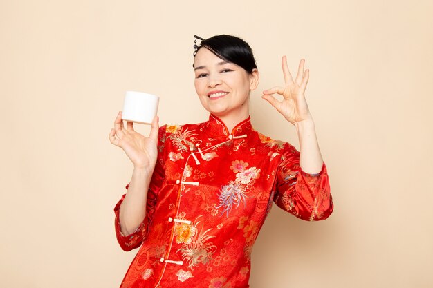 Una vista frontal hermosa geisha japonesa en el tradicional vestido rojo japonés con bastones para el pelo posando con crema puede sonreír en la ceremonia de fondo crema Japón