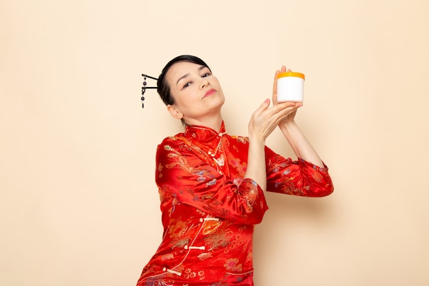 Foto gratuita una vista frontal hermosa geisha japonesa en el tradicional vestido rojo japonés con palitos de pelo posando sosteniendo la lata de crema en la ceremonia de fondo crema japón