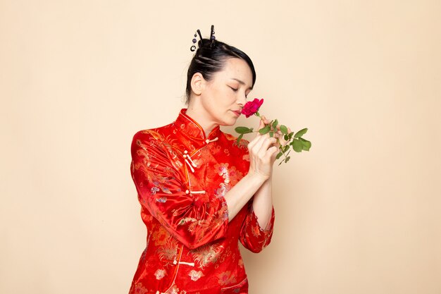 Foto gratuita una vista frontal hermosa geisha japonesa en el tradicional vestido rojo japonés con palos para el cabello con olor a rosa roja elegante en la ceremonia de fondo crema entretenido japón oriental