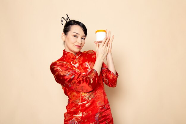 Una vista frontal hermosa geisha japonesa en el tradicional vestido rojo japonés con palos para el cabello posando sosteniendo la crema puede sonreír en la ceremonia de fondo crema Japón