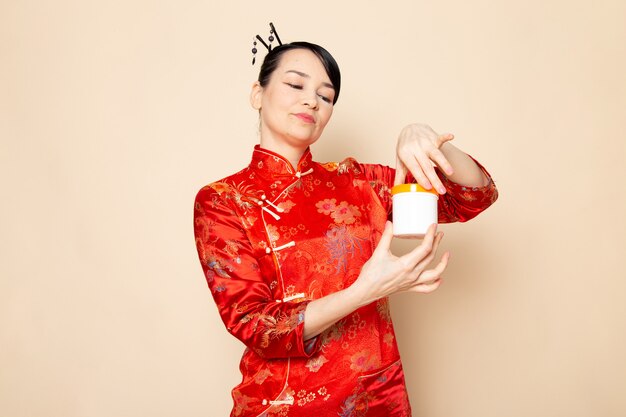 Una vista frontal hermosa geisha japonesa en el tradicional vestido rojo japonés con palos para el cabello posando sosteniendo la crema puede sonreír en la ceremonia de fondo crema Japón