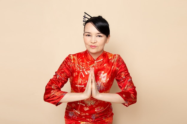 Una vista frontal hermosa geisha japonesa en el tradicional vestido rojo japonés con palos para el cabello posando con sus manos inclinándose sobre la ceremonia de fondo crema entretenida Japón oriental