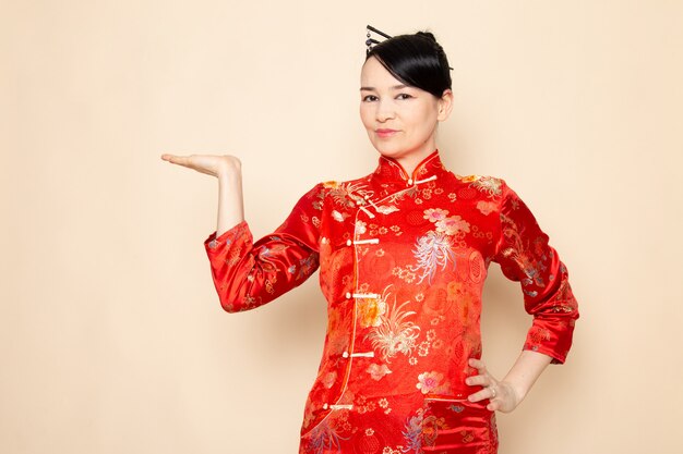 Una vista frontal hermosa geisha japonesa en el tradicional vestido rojo japonés con palos de pelo posando con las manos de pie sobre la ceremonia de fondo crema entretenido Japón oriental